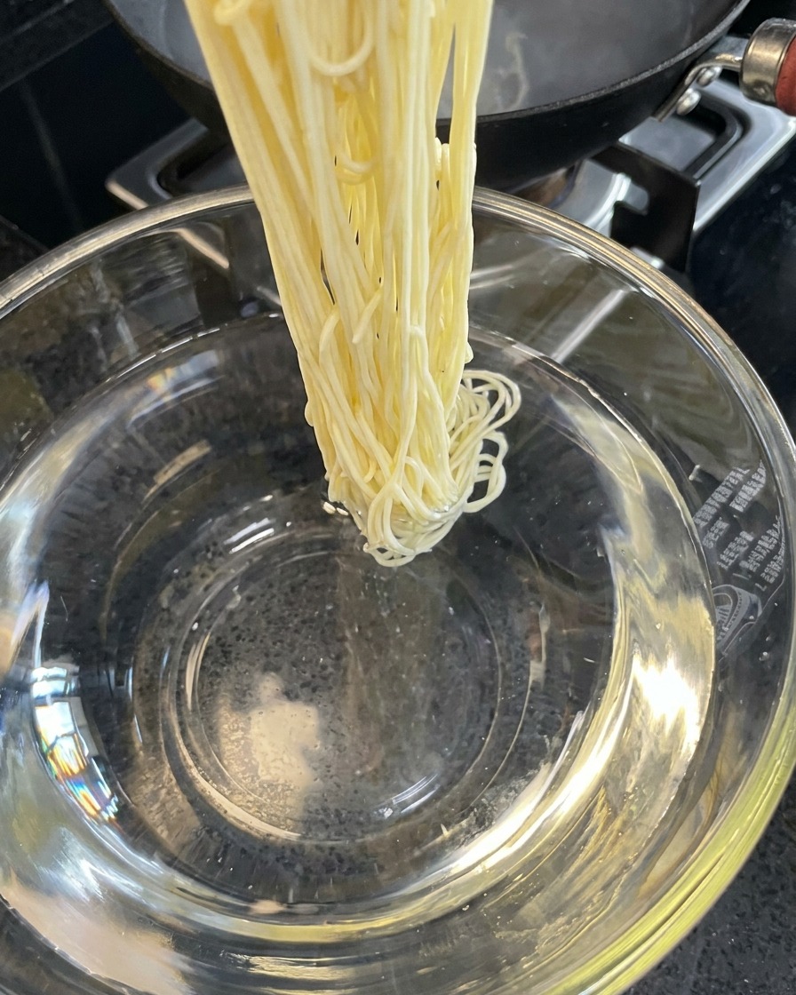 Cooked noodles being rinsed in a bowl of cold water.