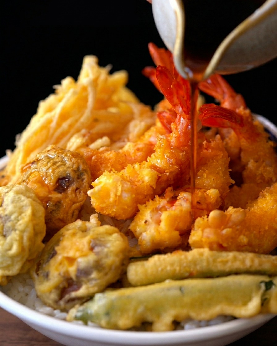 A dark, savory sauce being poured from a ceramic pitcher over a bowl of rice topped with crispy fried shrimp and vegetables.