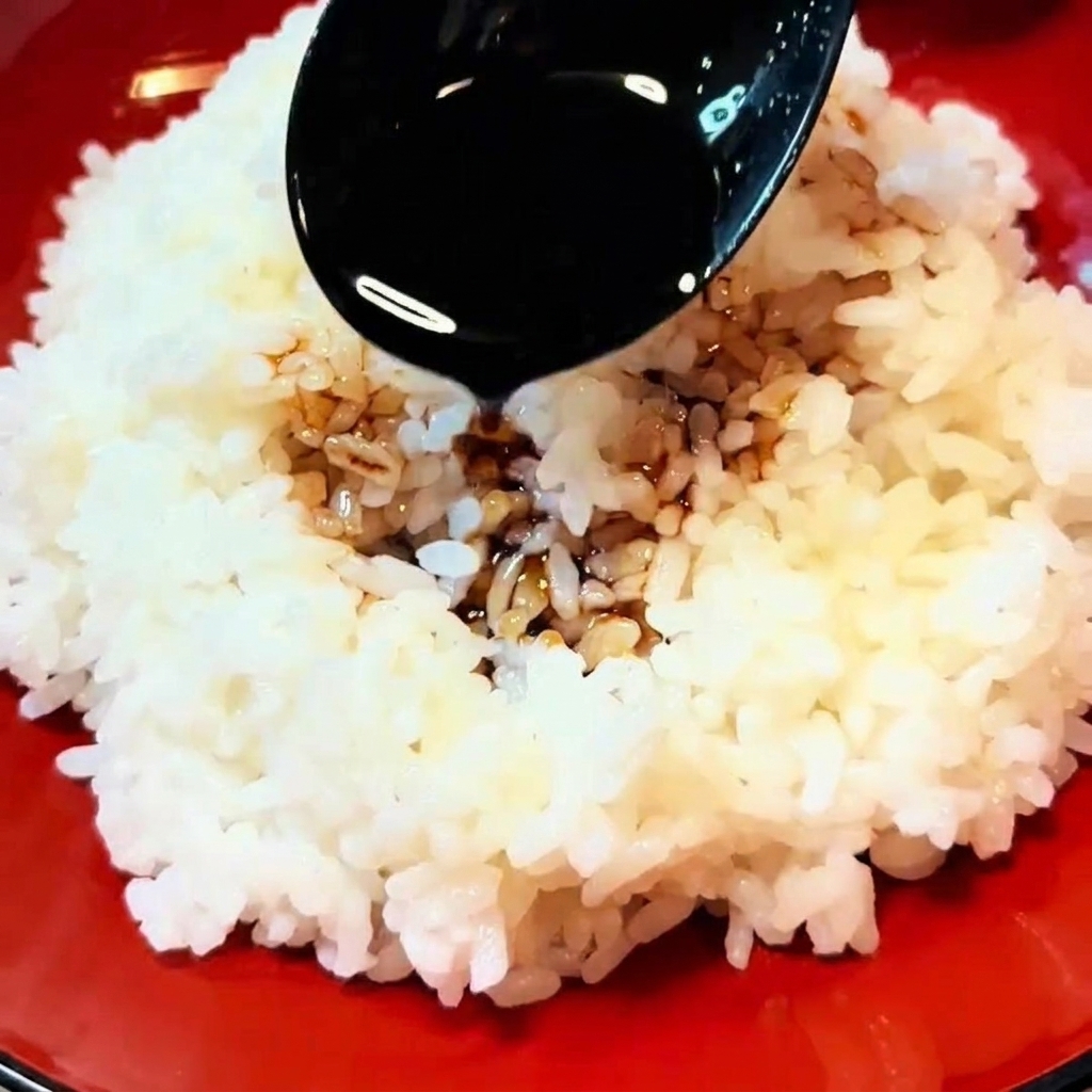 A black spoon drizzling dark brown sauce over a bowl filled with steaming white rice.