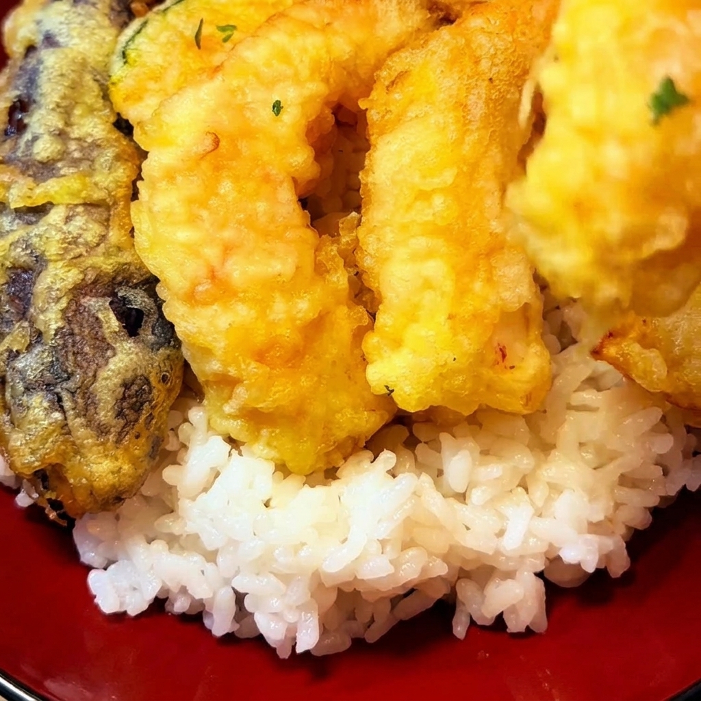 Golden-brown fried tempura pieces, including shrimp and vegetables, being placed on top of a bed of white rice in a red bowl.