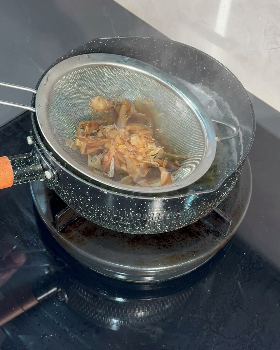 A fine-mesh metal strainer filled with bonito flakes being dipped into a pot of simmering water.