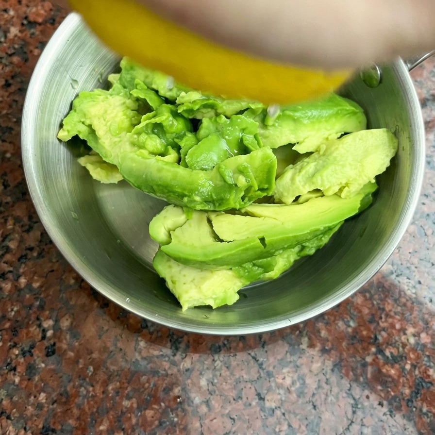 Fresh lemon juice being squeezed by hand onto halved avocados inside a small stainless steel bowl.