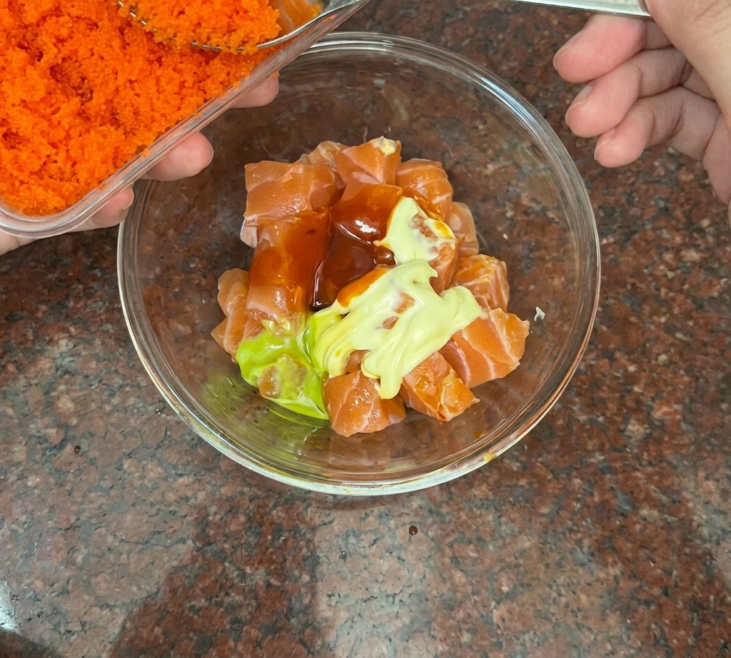 A silver spoon adding bright orange fish roe to a bowl of cubed salmon.