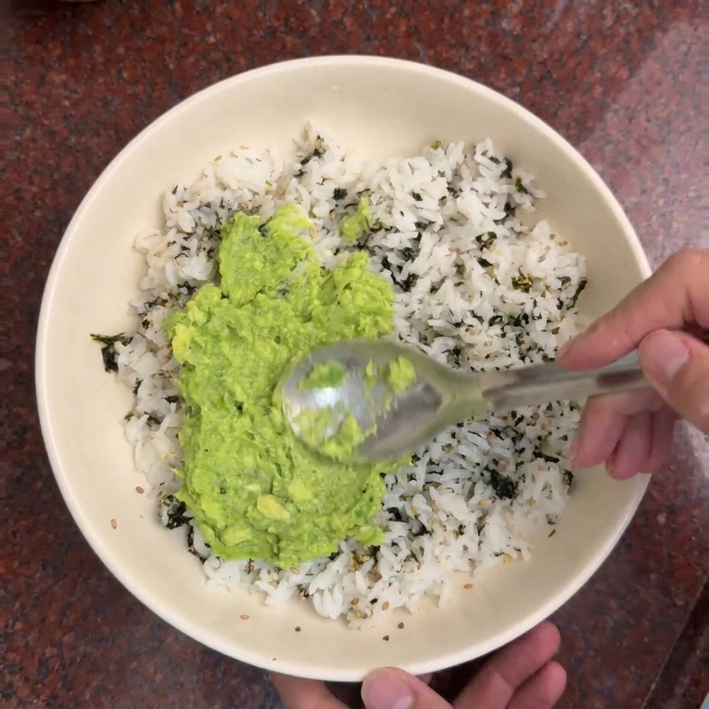 A spoon spreading bright green mashed avocado over a bowl of white rice seasoned with dark seaweed strips.