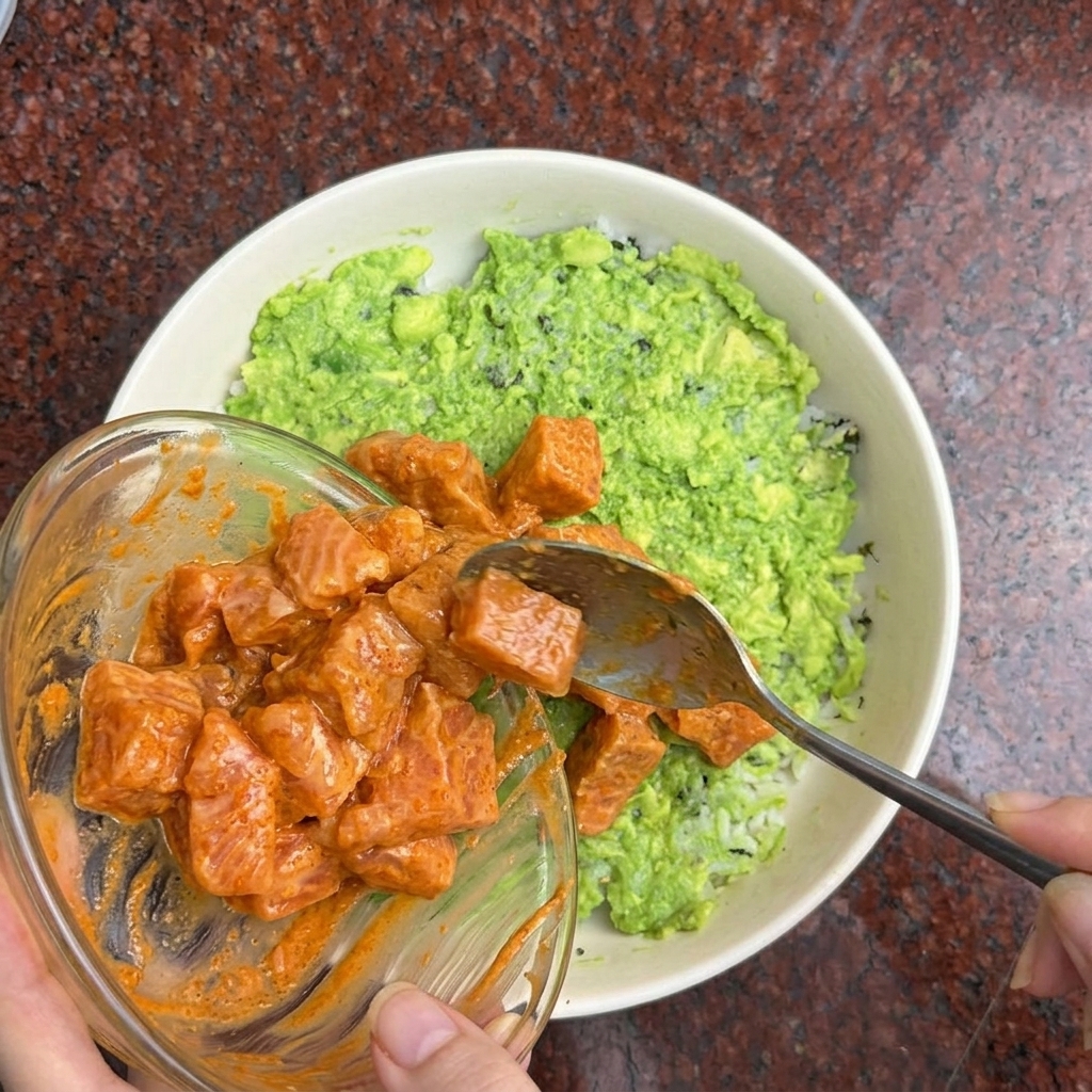 A hand using a spoon to place orange-marinated salmon cubes on top of a rice and avocado bowl.
