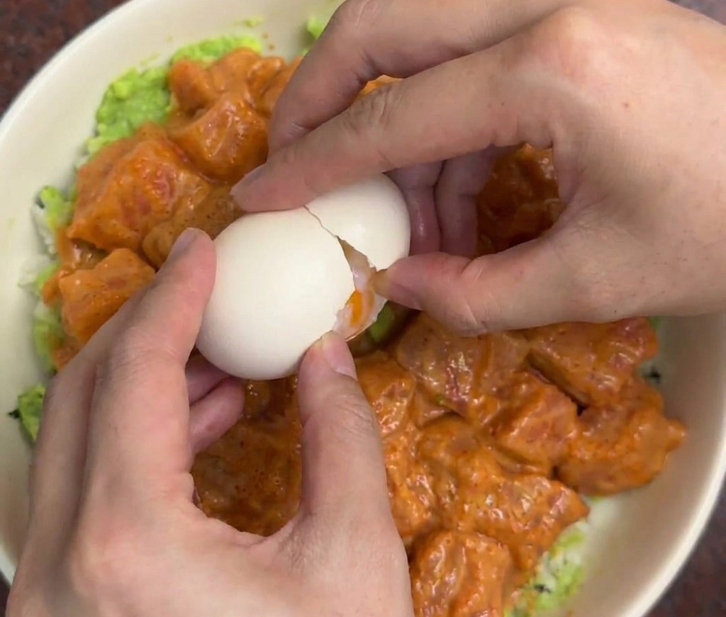Hands gently cracking a white onsen egg into the center of a salmon and avocado rice bowl.