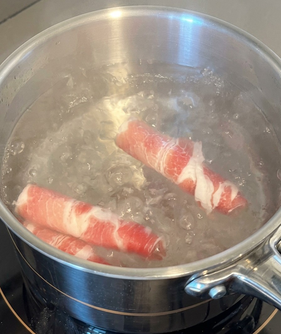 Two rolls of raw fatty beef slices being submerged in a stainless steel pot of boiling water.