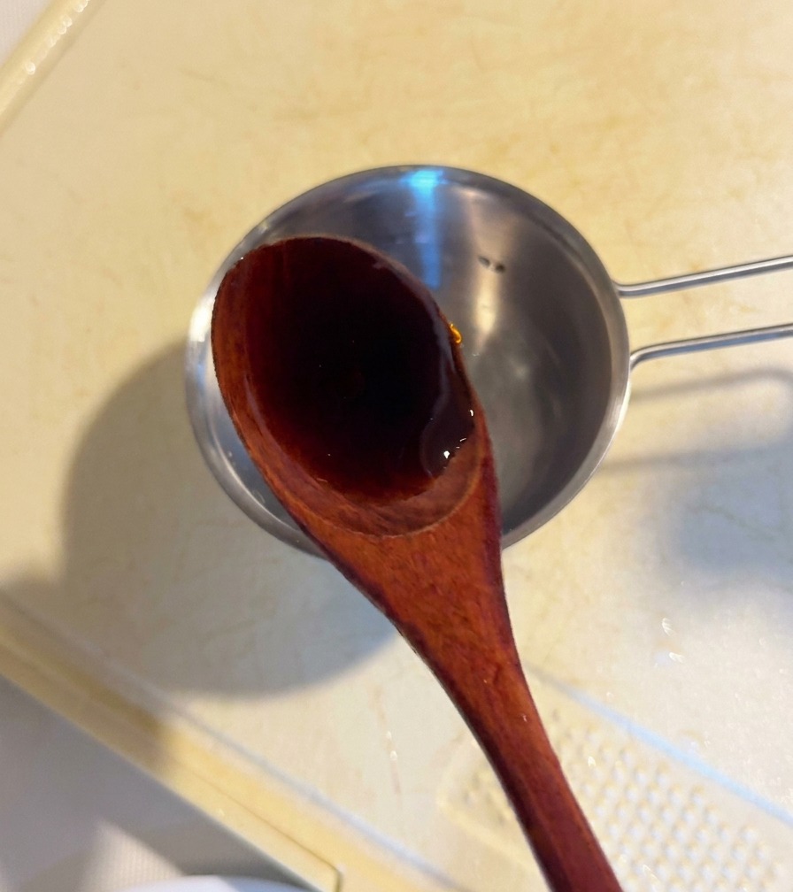 A wooden spoon pouring dark oyster sauce into a small metal prep bowl containing sauce ingredients.