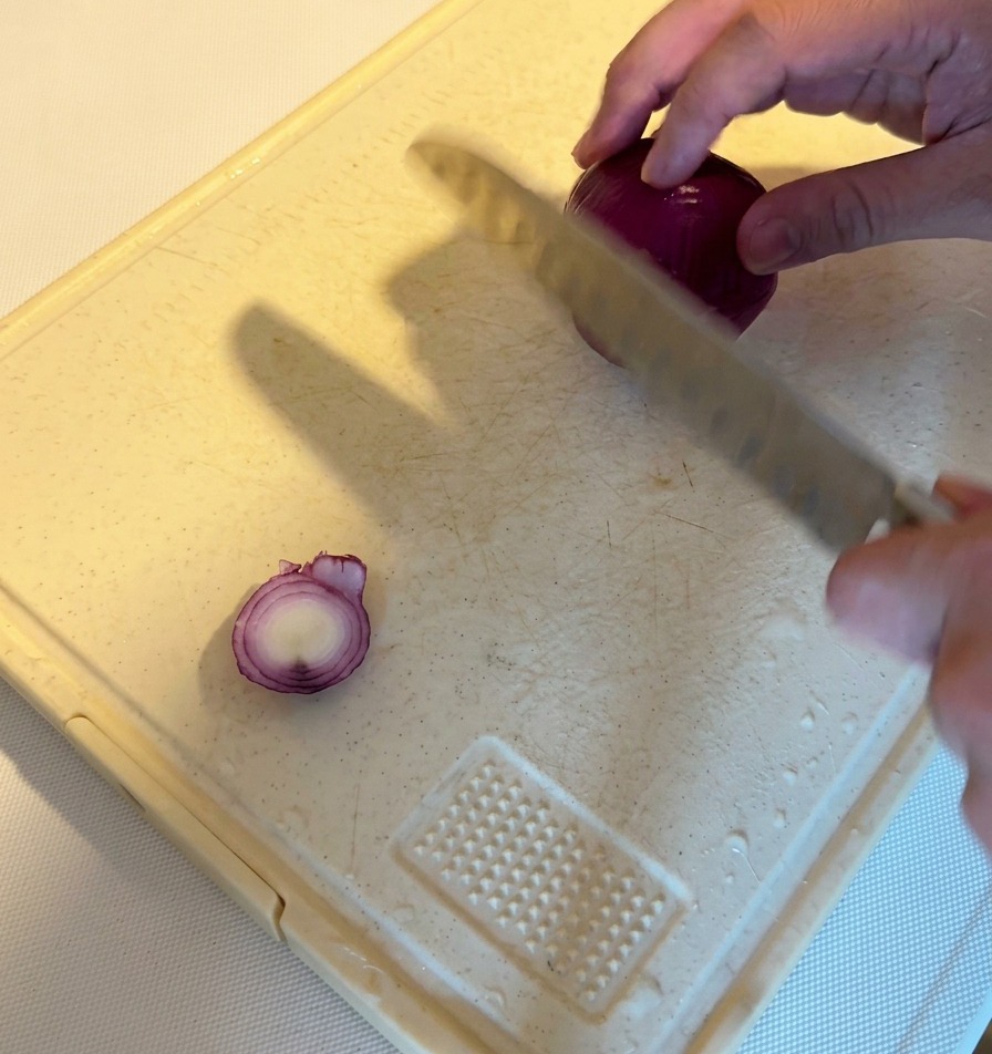 A person using a chefs knife to slice a whole red onion into thin wedges on a white cutting board.