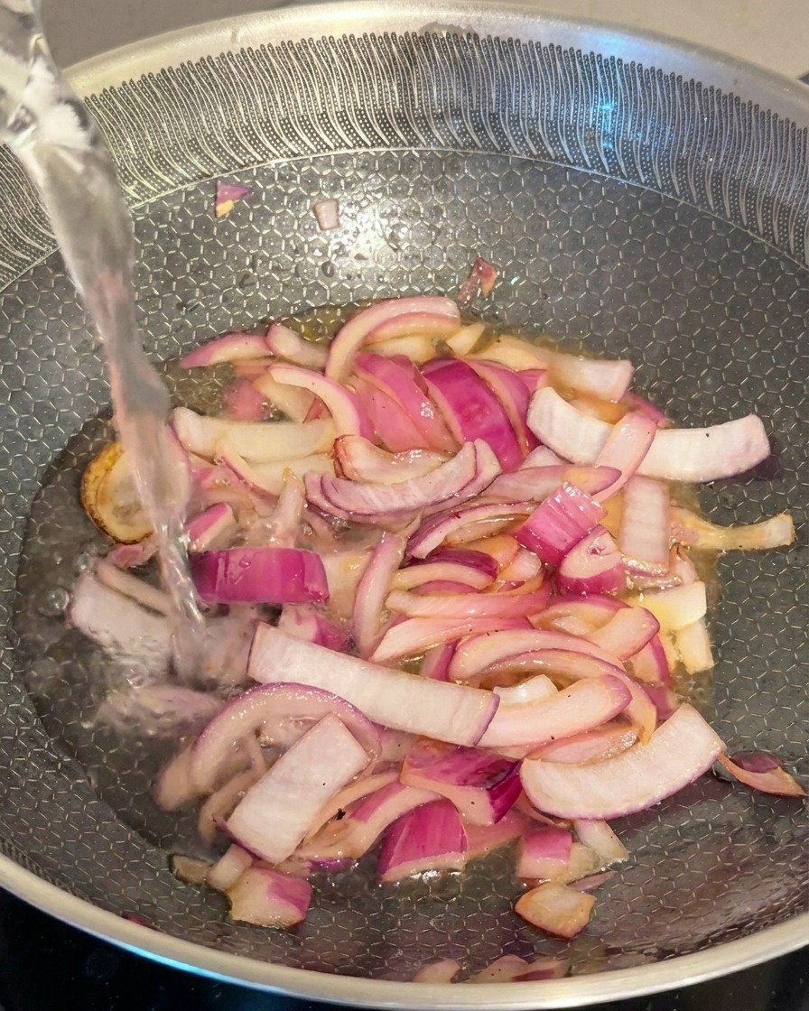 A stream of clear water being added to a pan of stir-fried red onions.