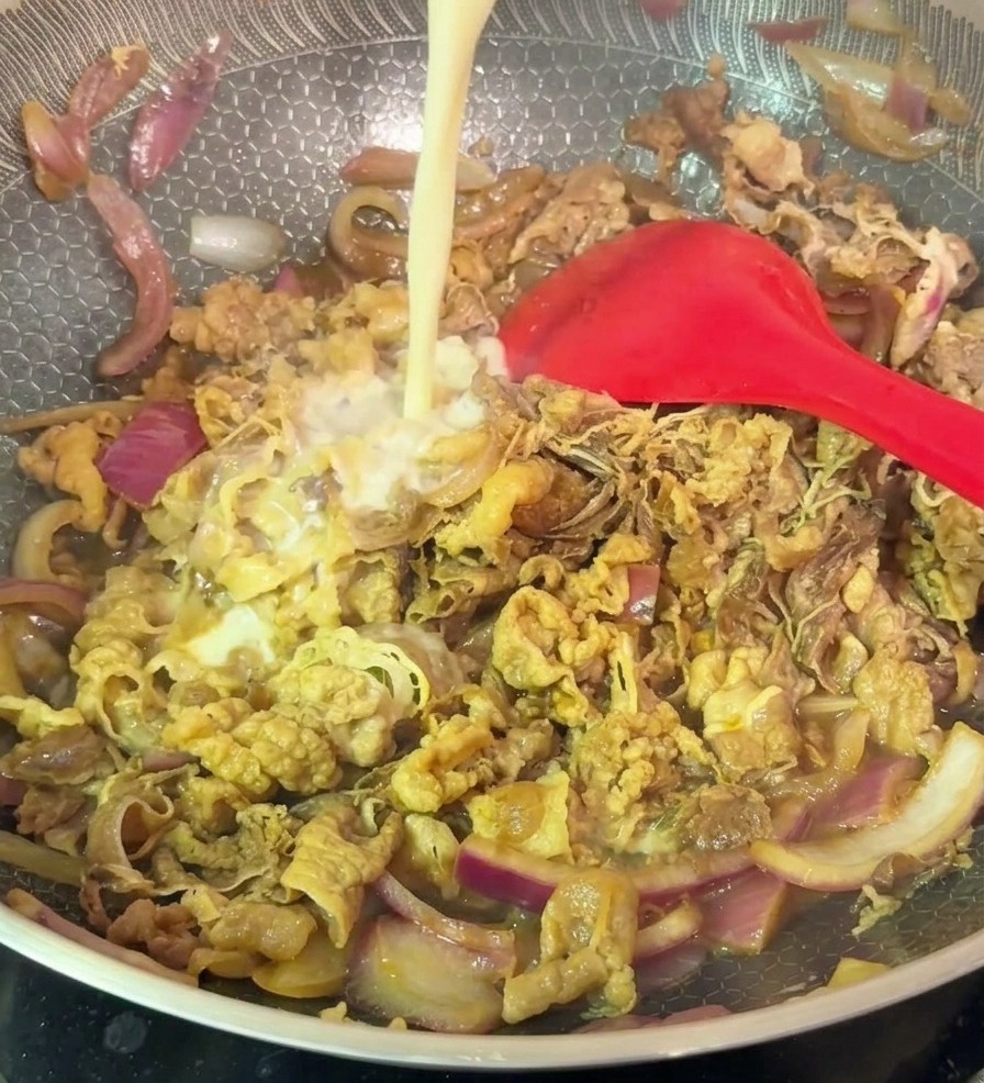 A stream of pale starch slurry being poured into a hexagonal-patterned wok filled with stir-fried beef and red onions.