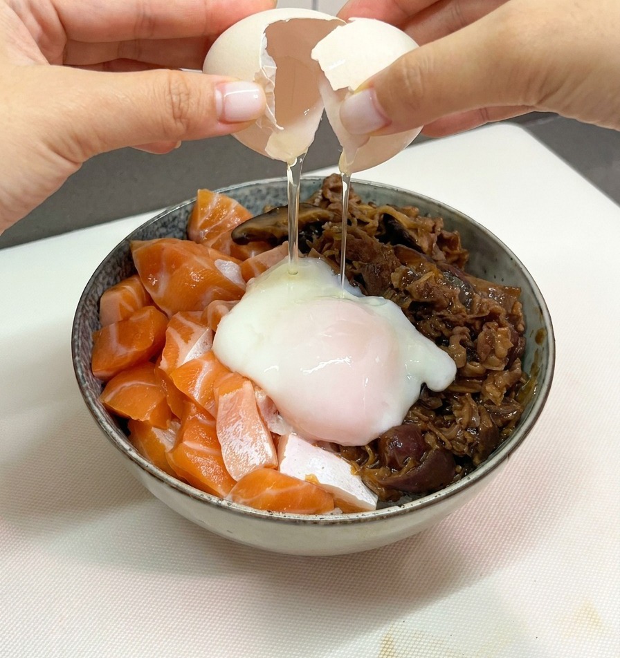 Two hands carefully cracking a white egg shell to release a soft jiggly onsen egg onto a bowl of salmon and beef rice.