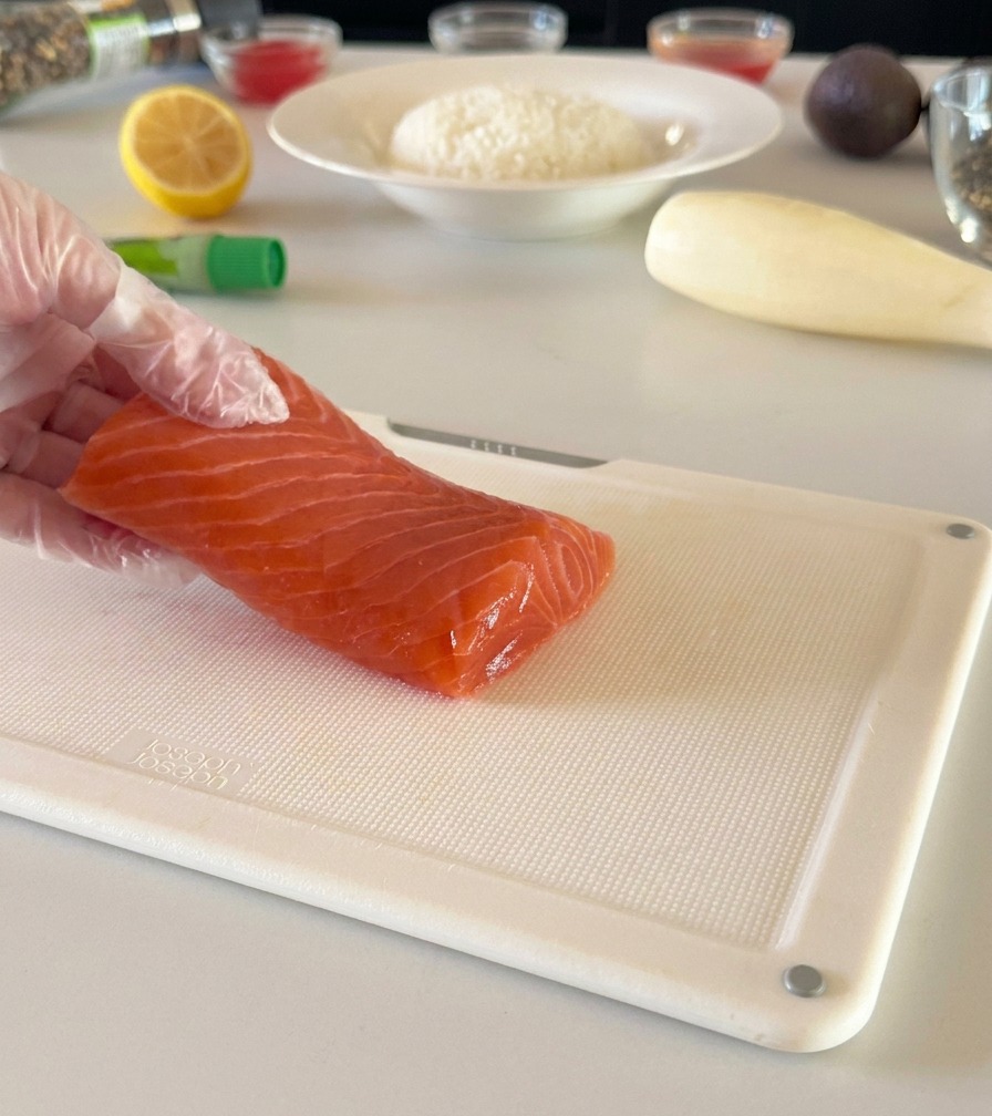 A gloved hand holding a fresh salmon fillet on a white cutting board, preparing to slice it into cubes.
