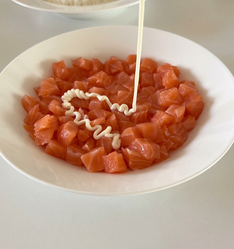 Creamy white mayonnaise being drizzled from a bottle onto a bowl of fresh, orange salmon cubes.