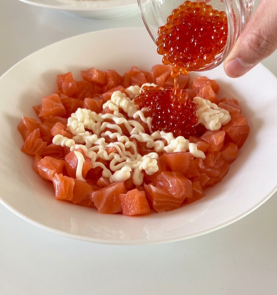 Freshly cubed salmon in a white bowl being topped with bright red fish roe and creamy mayonnaise.