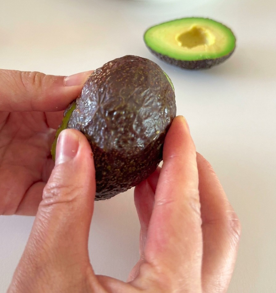 Two hands carefully peeling the dark skin off a ripe, green avocado half.