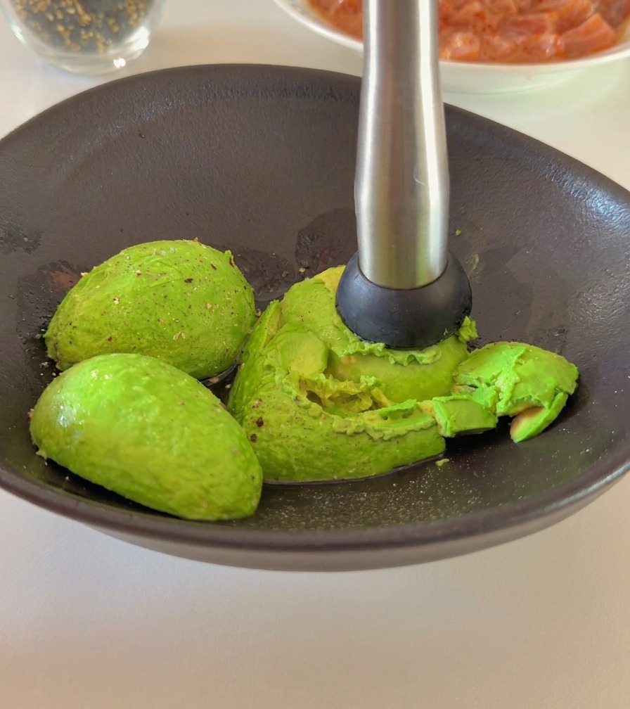 A person using a metal hand masher to crush ripe avocado halves in a dark ceramic bowl.