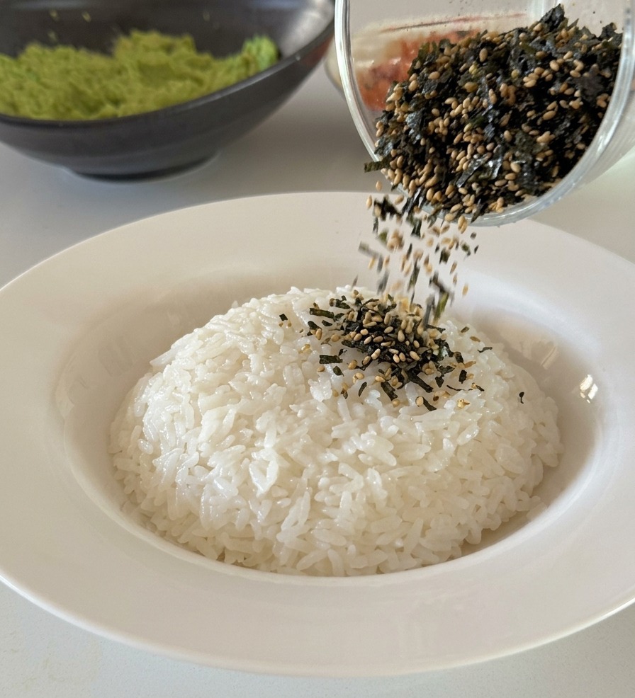 A mixture of black seaweed strips and white sesame seeds being poured from a glass cup onto a neat mound of white rice.