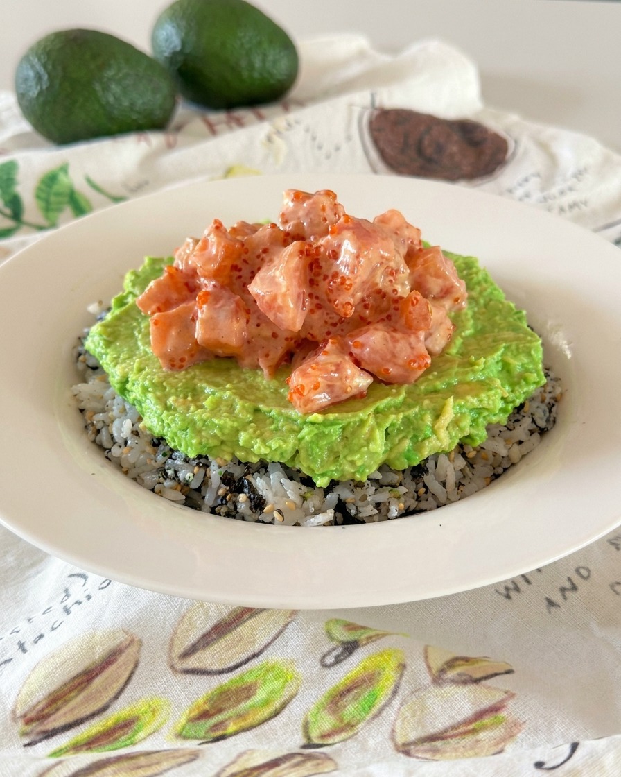 A poke bowl showing distinct layers of seasoned rice, mashed avocado, and creamy marinated salmon cubes.