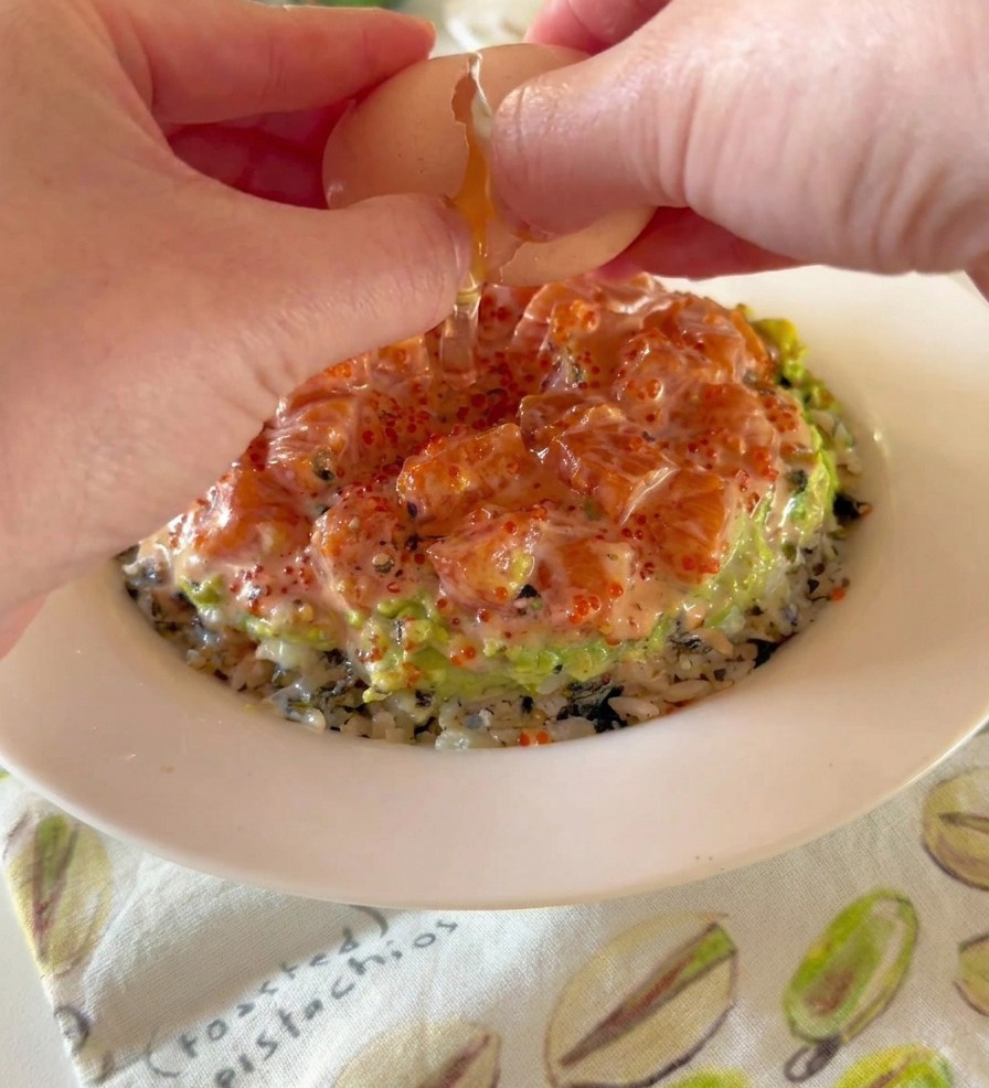 A persons hands cracking a soft-boiled onsen egg over a bowl layered with rice, mashed avocado, and cubed salmon.