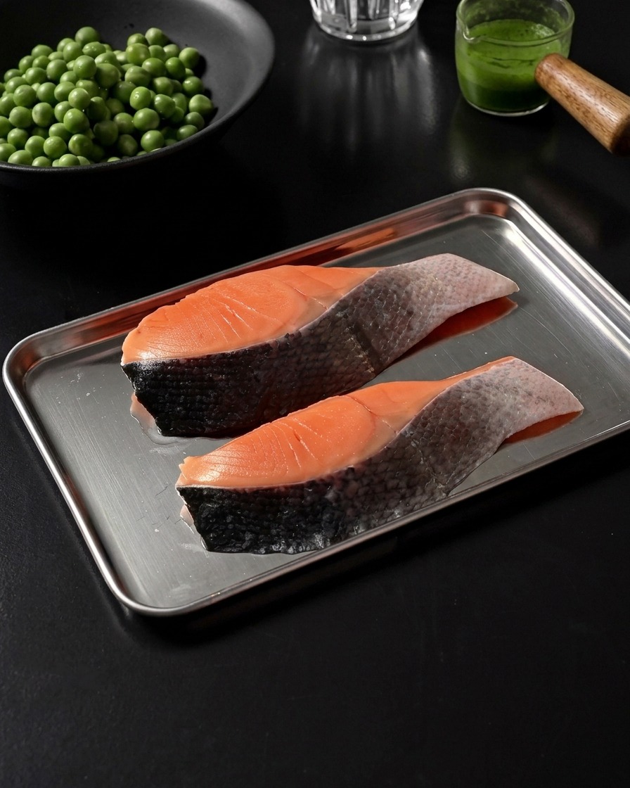 Two raw salmon fillets resting on a stainless steel tray, lightly seasoned with salt and black pepper.