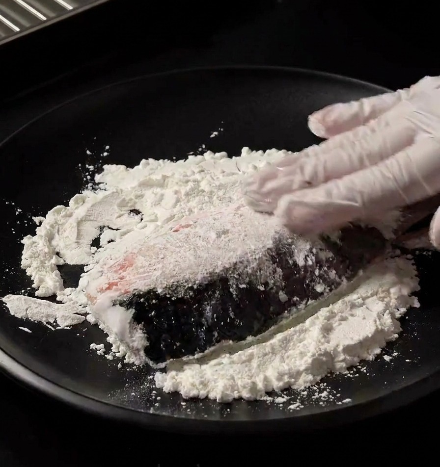 A gloved hand pressing a fresh salmon fillet into a bed of white starch on a black plate to coat it evenly.