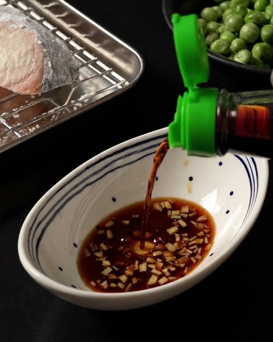 Dark soy sauce being poured into a white bowl containing a base of minced ginger and honey.