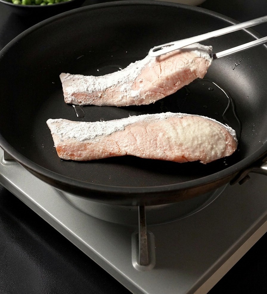 Metal tongs placing a starch-dredged salmon fillet into a hot pan alongside another fillet.
