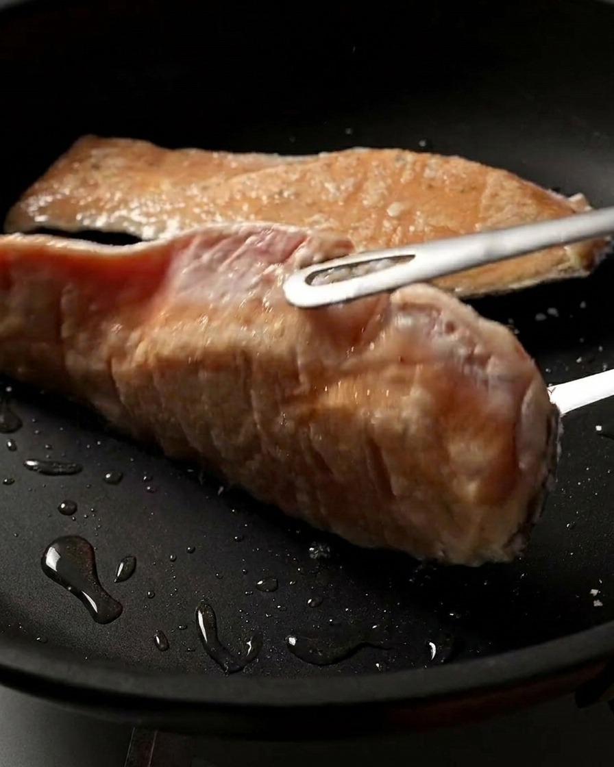 A close-up shot of a salmon fillet being flipped in the pan, showing its perfectly browned and crispy seared surface.