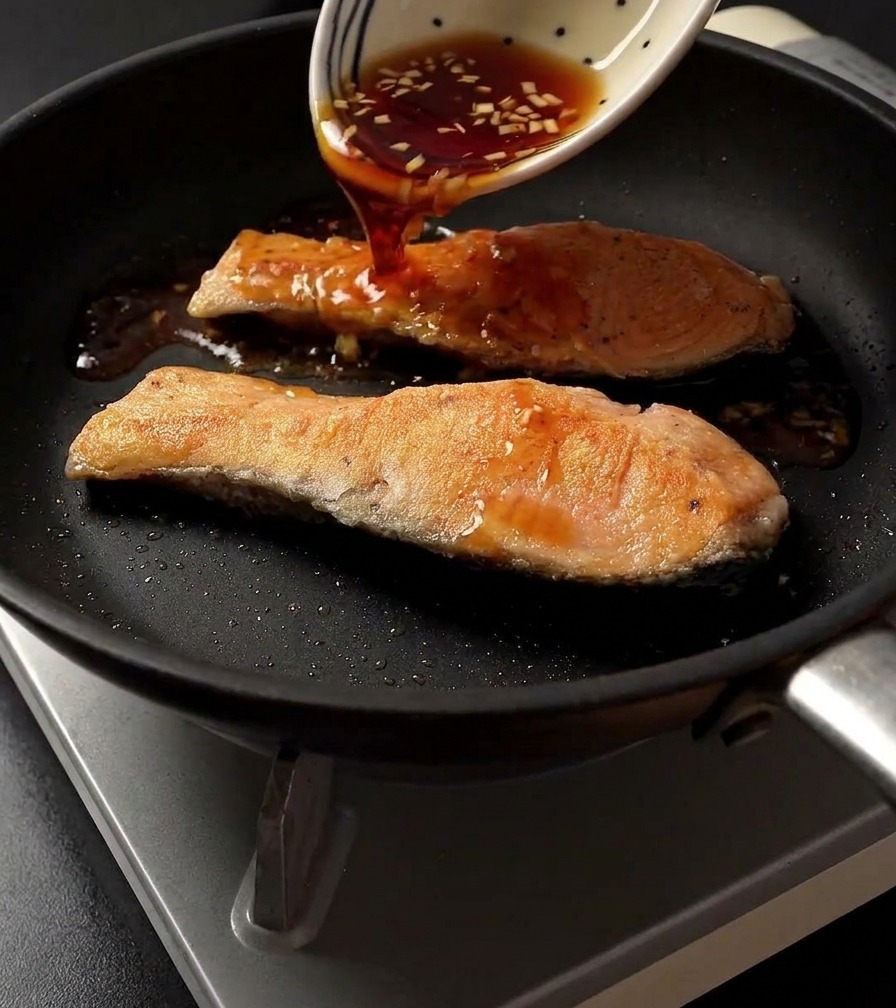 Pouring dark teriyaki sauce from a patterned white bowl onto two seared salmon fillets in a skillet.
