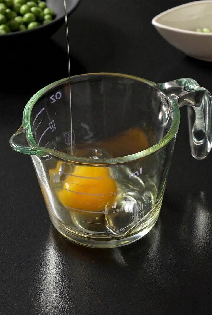 A fresh egg being cracked into a clear glass measuring cup, with a bowl of green peas in the blurred background.