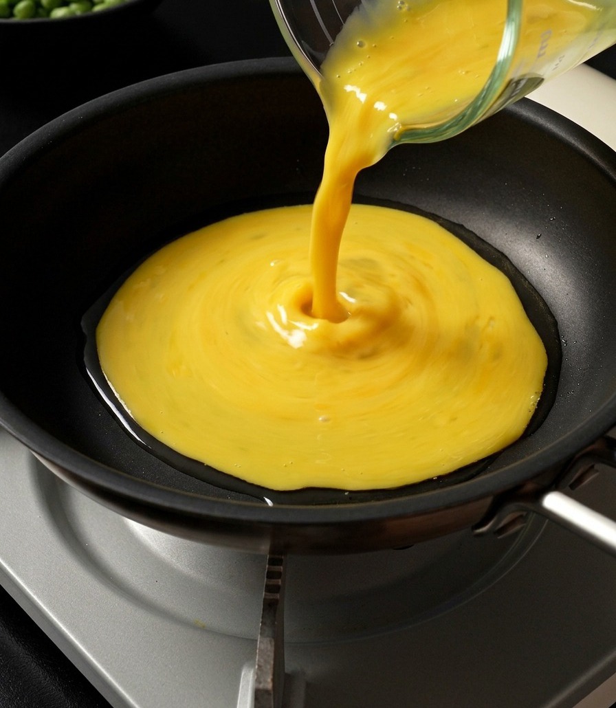 Beaten yellow egg mixture being poured from a glass measuring cup into a wide frying pan to form a thin omelet.