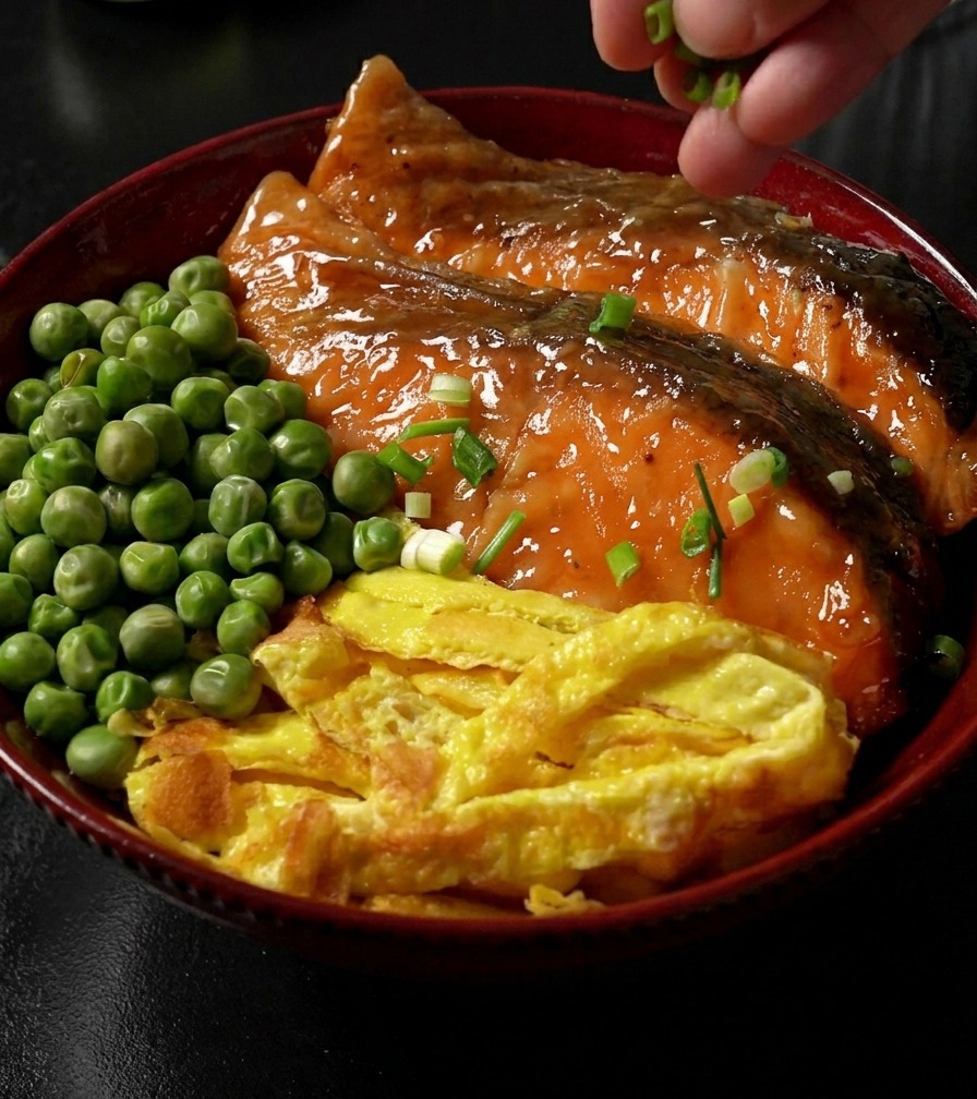 A hand sprinkling sliced green scallions over a completed rice bowl topped with teriyaki-glazed salmon, egg strips, and green peas.