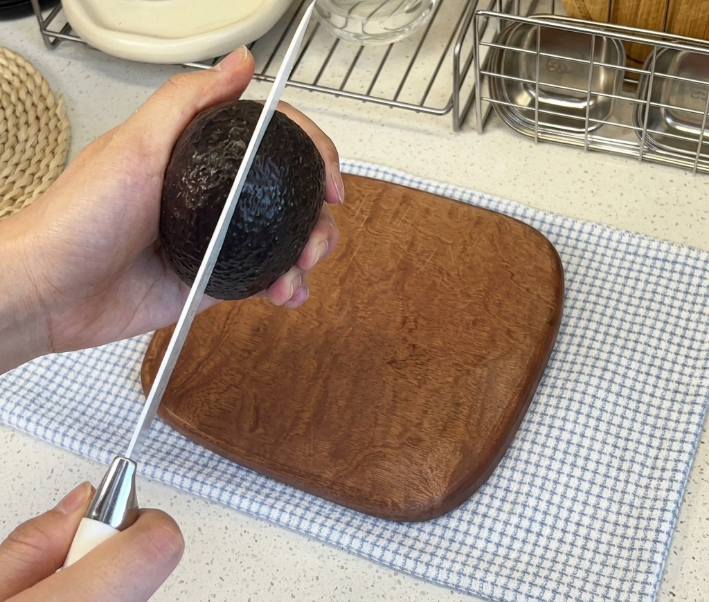 A persons hands holding a dark-skinned avocado while slicing it vertically with a sharp kitchen knife.