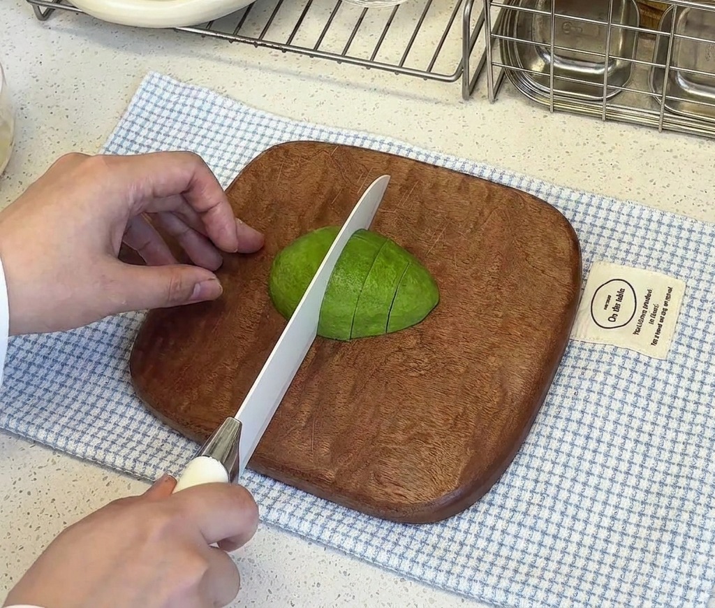 A hand steadying a peeled avocado half on a wooden board while a white knife cuts it into small cubes.