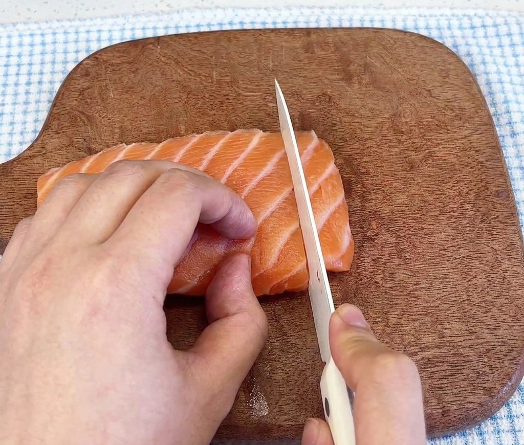 Close-up of a sharp knife slicing through a fresh, marbled salmon fillet on a dark wood cutting board.