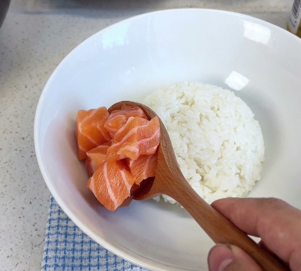 A wooden spoon lifting a pile of fresh salmon cubes and placing them into a white bowl of steamed rice.