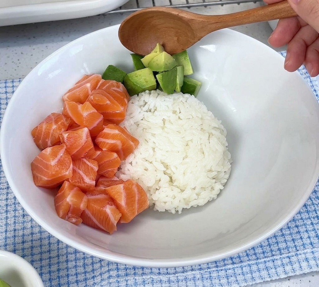 Diced avocado being spooned into a white bowl next to a pile of raw salmon cubes over rice.