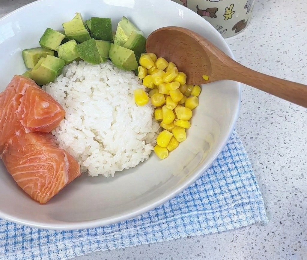 A wooden spoon adding bright yellow corn kernels into a white bowl filled with rice, diced avocado, and fresh salmon pieces.