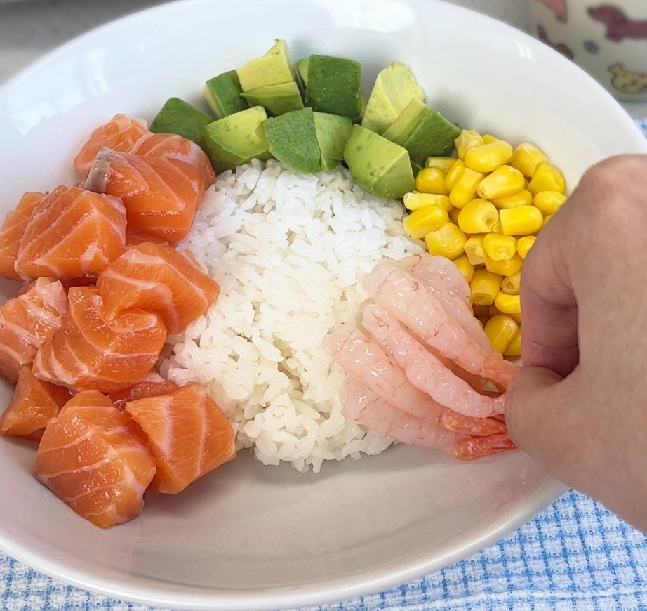 A hand placing pink, translucent sweet shrimp onto the rice in a bowl alongside salmon and corn.