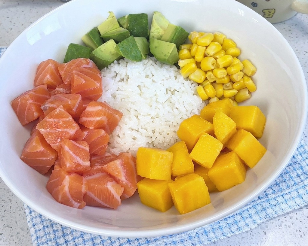 A colorful poke bowl with white rice, salmon, avocado, corn, and shrimp, now featuring a pile of bright yellow mango cubes.