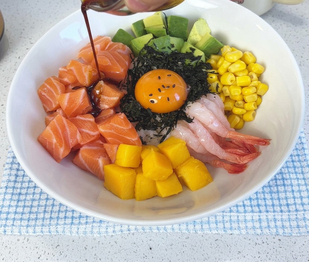 Dark wasabi soy sauce being poured from a small glass bowl over the assembled salmon poke bowl and egg yolk.