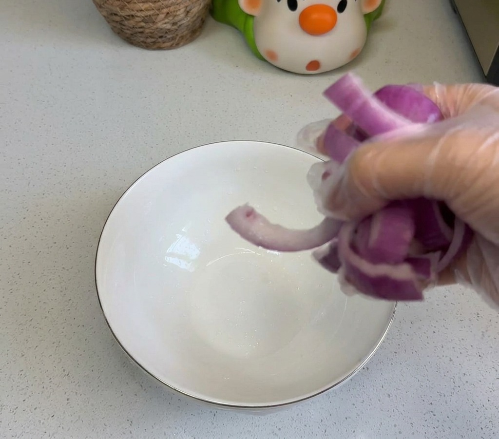 A person adding sliced red onions into a white ceramic prep bowl on a clean kitchen counter.