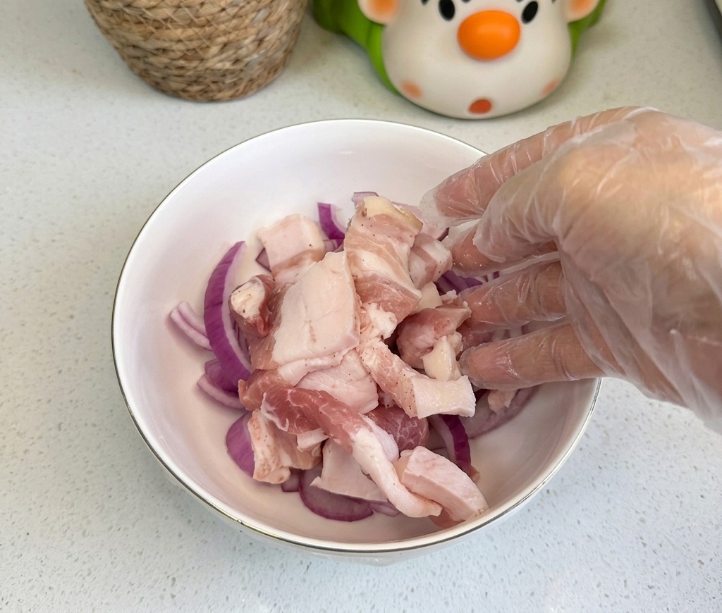 Raw pork belly slices being added to a white bowl containing thinly sliced red onions.