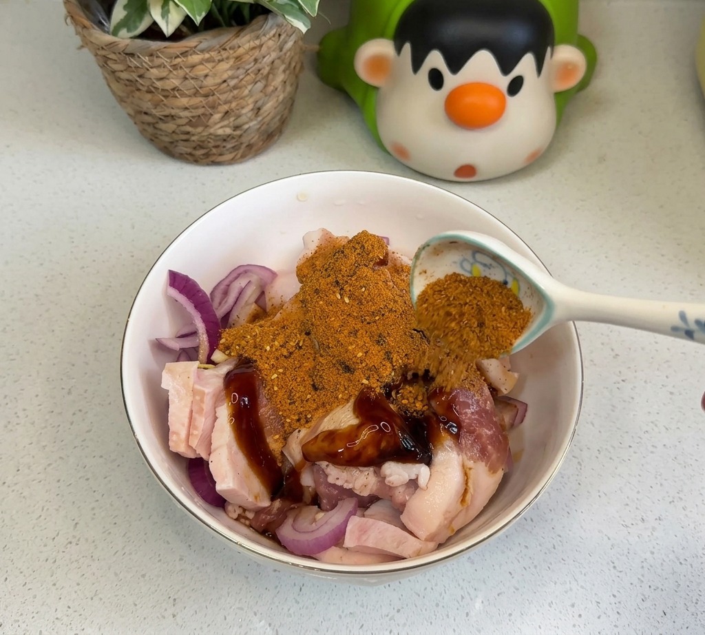 A spoonful of orange BBQ seasoning and red chili powder being sprinkled over the marinated pork belly and onions.