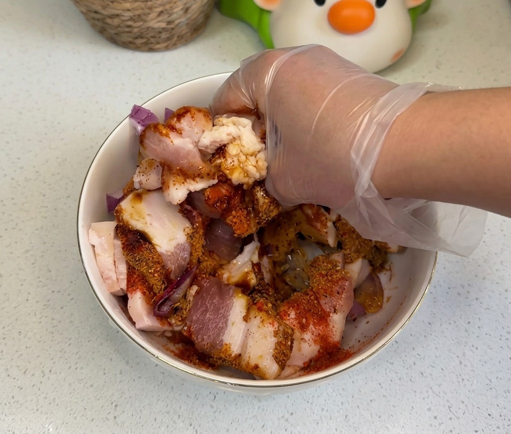 A hand in a clear plastic glove thoroughly mixing raw pork belly slices with onions and colorful seasonings in a bowl.