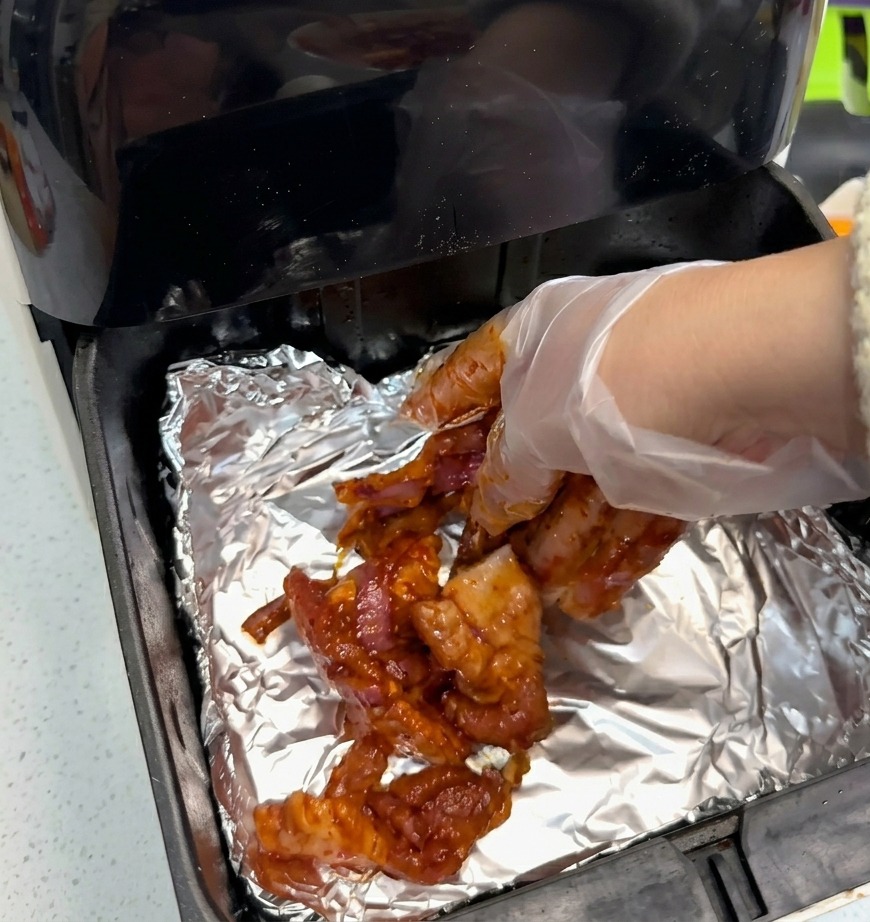 A hand wearing a clear plastic glove placing red-marinated pork belly slices onto silver aluminum foil inside a black air fryer basket.