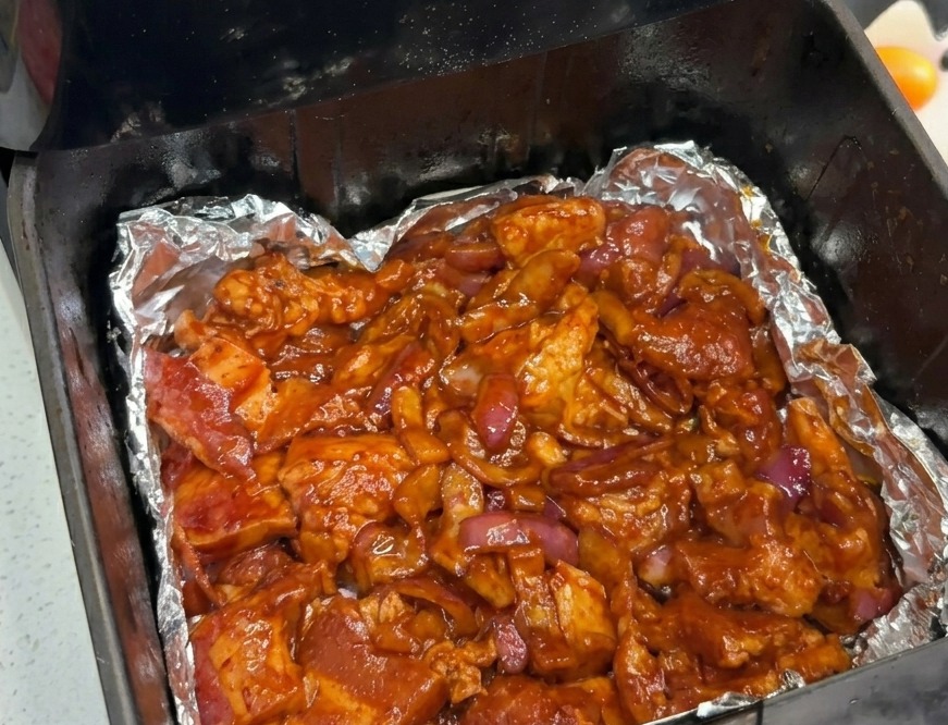 Marinated pork belly and red onions spread out evenly across the bottom of an air fryer basket lined with foil.
