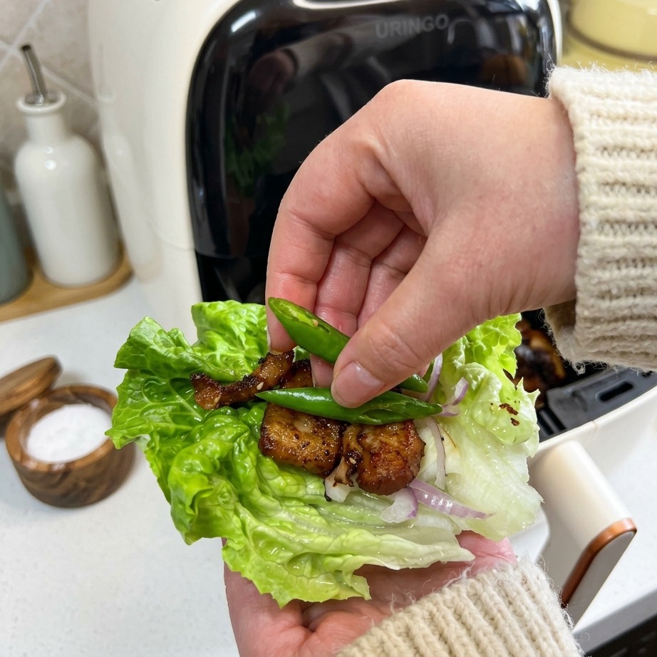A hand placing two small, bright green chili peppers onto a lettuce wrap containing crispy pork belly and onions.