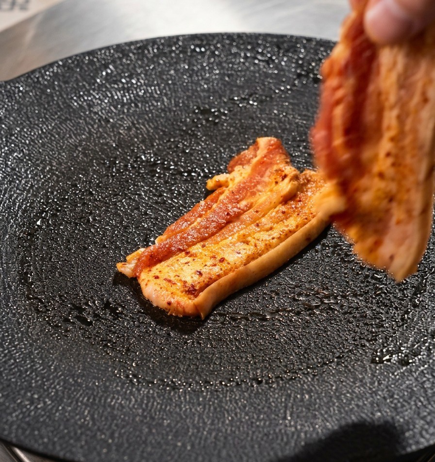 A hand placing a strip of marinated pork belly onto a textured black round grill pan.